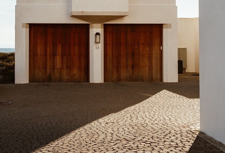 Two wooden single-car garage doors and cobblestone driveway outside beige home