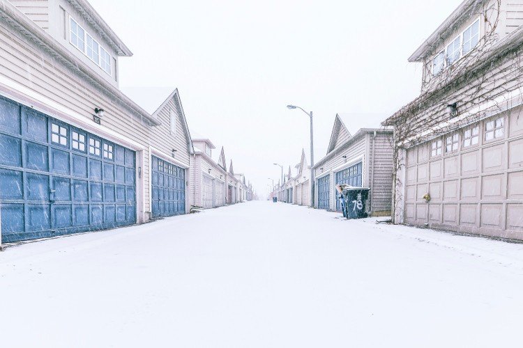 Two-story houses with blue and tan-colored garage doors lining both sides of a residential street on snowy day